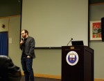 Ben Pender-Cudlip stands with a microphone in front of projection screen, to the left of a podium with the SUNY Ulster logo